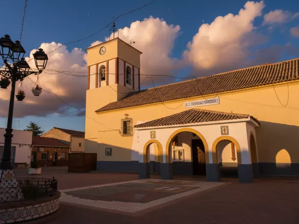 Plaza de un pueblo al atardecer con una iglesia de paredes amarillas y un arco de entrada bajo un cielo con nubes.
