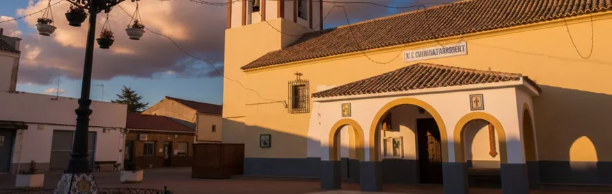 Plaza de un pueblo al atardecer con una iglesia de paredes amarillas y un arco de entrada bajo un cielo con nubes.