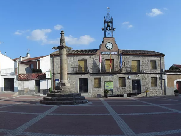 Una amplia plaza pavimentada que alberga un monumento de piedra central y un edificio consistorial con un reloj y campanas en la parte superior.