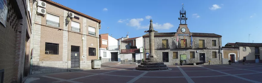 Una amplia plaza pavimentada que alberga un monumento de piedra central y un edificio consistorial con un reloj y campanas en la parte superior.