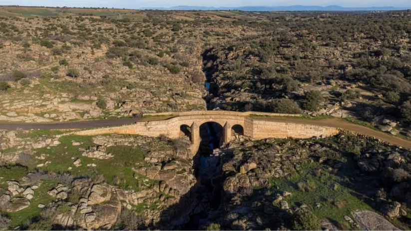 Fachada de una pequeña construcción de ladrillo y piedra con una espadaña sencilla, una campana y una palmera en primer plano.