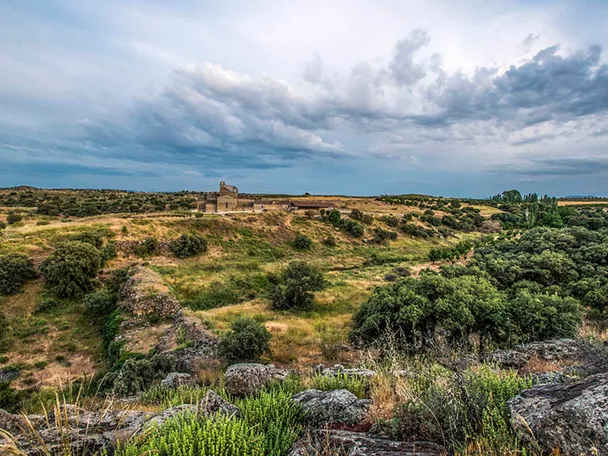 Paisaje natural con colinas, vegetación baja y edificio de piedra en la distancia