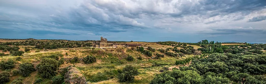 Paisaje natural con colinas, vegetación baja y edificio de piedra en la distancia