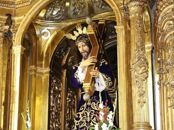 Escultura religiosa con cruz en retablo dorado y flores blancas.