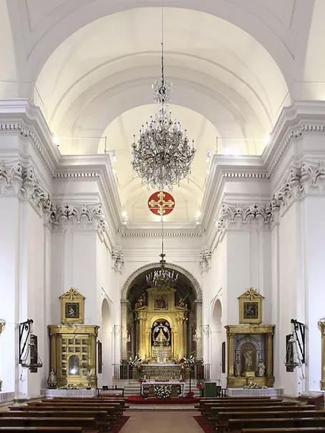 Nave central de iglesia con bóveda blanca, candelabro y retablo iluminado.