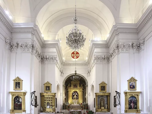 Nave central de iglesia con bóveda blanca, candelabro y retablo iluminado.