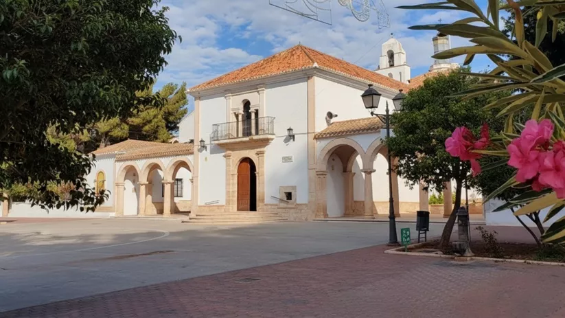 Plaza con edificio blanco porticado, tejados de teja y flores rosadas en primer plano.
