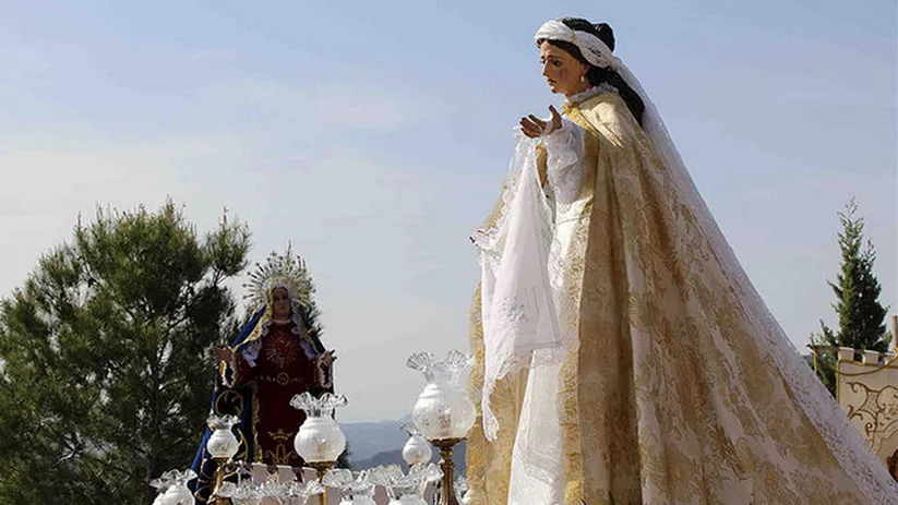Imagen religiosa femenina vestida de blanco y dorado sobre un paso procesional al aire libre.