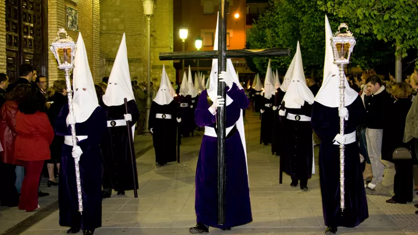 Nazarenos con túnicas oscuras portando cruces y faroles en procesión nocturna