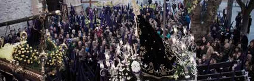 Multitud acompañando un paso procesional decorado con flores y cirios en la calle.