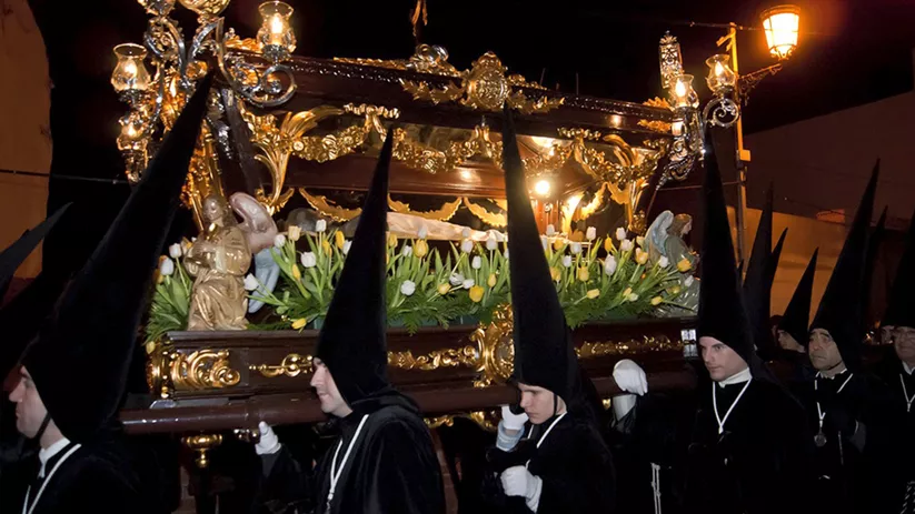 Paso procesional nocturno con figuras religiosas y flores, escoltado por nazarenos con capirotes negros.