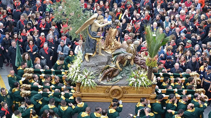 Paso procesional con figuras religiosas rodeado de portadores y público en una plaza.