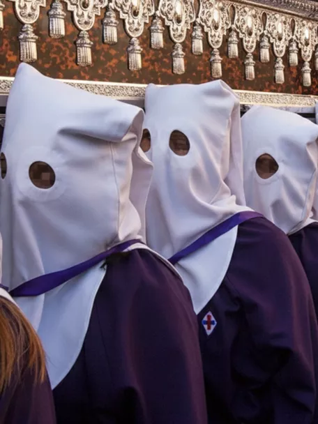 Fila de penitentes con túnicas moradas y capirotes blancos durante una procesión.