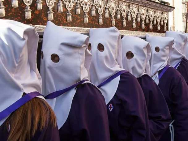 Fila de penitentes con túnicas moradas y capirotes blancos durante una procesión.