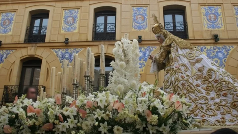 Paso con figura religiosa y manto bordado, rodeado de flores, frente a una fachada con azulejos.