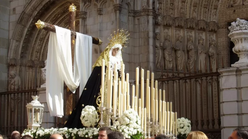Imagen religiosa con figura mariana, cruz y velas frente a una portada gótica, rodeada de flores blancas y público.