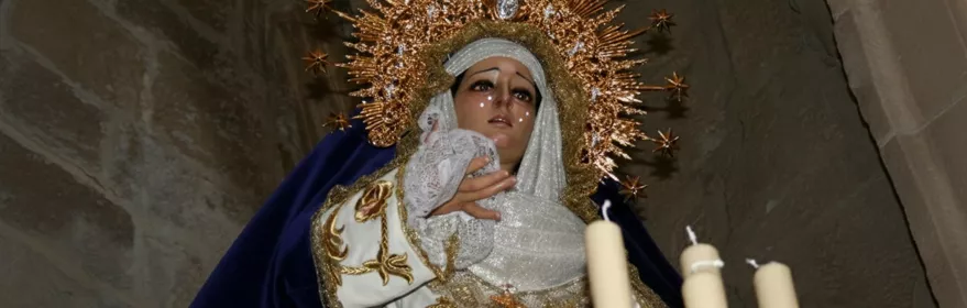 Imagen de una figura religiosa con corona dorada y manto azul, junto a velas encendidas en interior de piedra.