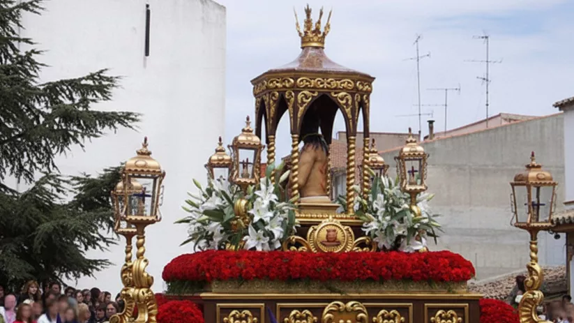Paso procesional dorado con flores rojas y faroles, rodeado de personas y penitentes con capirote morado.