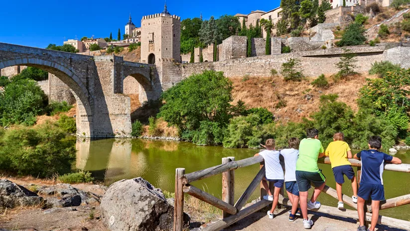 Puente de piedra medieval sobre un río con murallas al fondo.