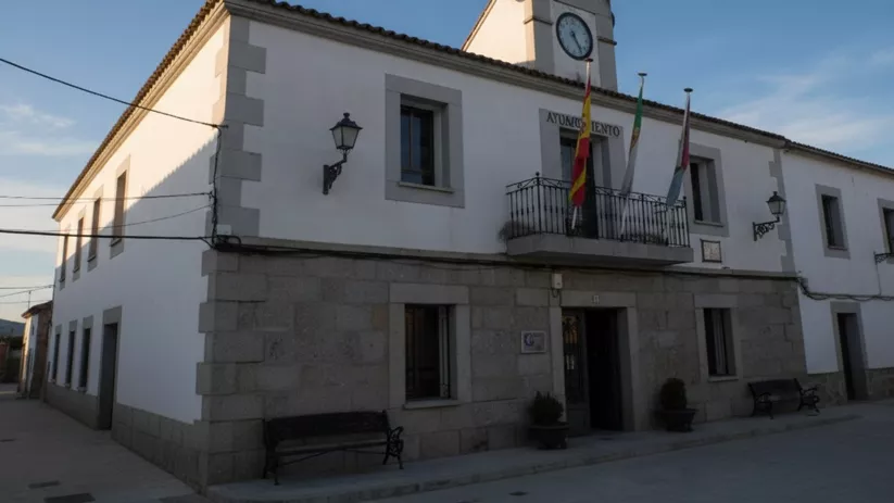 Fachada de un edificio municipal de dos plantas, pintado de blanco con la base de piedra, que cuenta con balcones de hierro y un reloj en la parte superior.