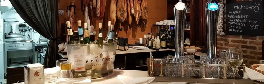 Barra de taberna con copas colgadas, vinos en cubitera y jamones al fondo.