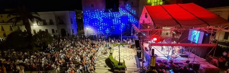 Concierto nocturno en plaza con escenario iluminado y público de pie.