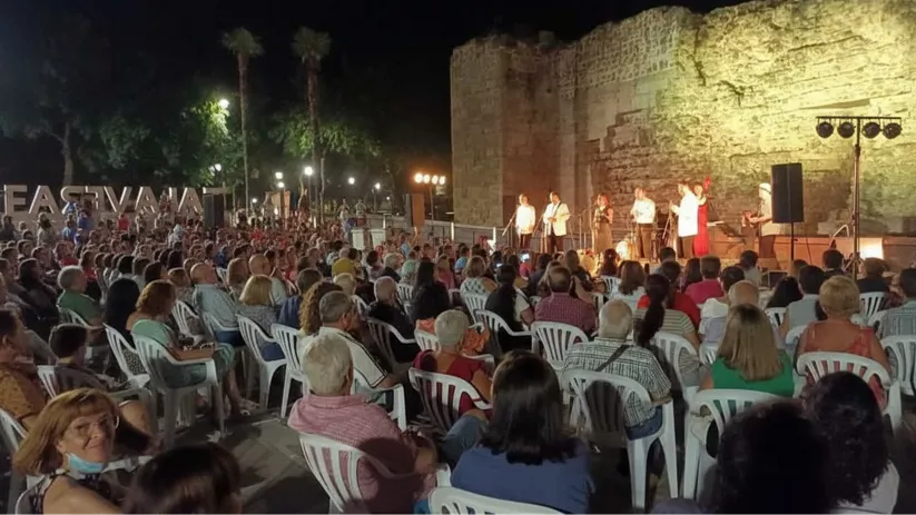 Concierto al aire libre con escenario iluminado junto a muralla histórica y público sentado.