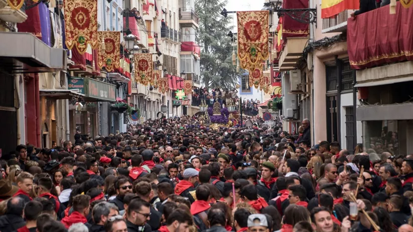 Calle estrecha abarrotada con estandartes colgantes y una procesión al fondo..