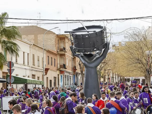 Personas vestidas de morado se concentran junto a escultura de tambor gigante.