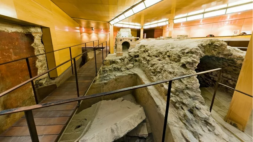 Pasarela interior junto a excavación y estructuras antiguas, con paredes de madera y luz artificial.