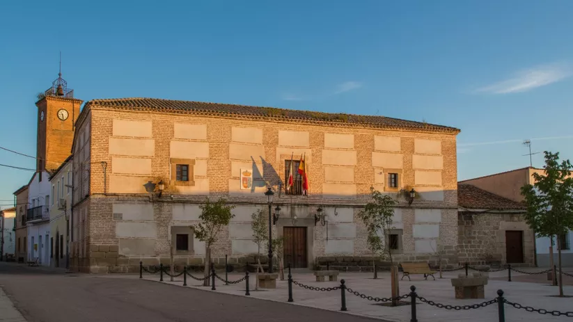 Fachada de un edificio civil de dos plantas con muros de ladrillo y piedra, situado frente a una plaza con bancos y árboles jóvenes, con una torre de reloj al fondo.