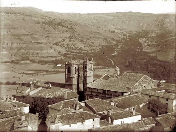 Fotografía histórica de un casco urbano con tejados tradicionales y torres al fondo.