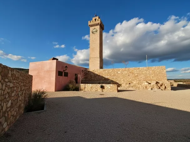 Torre de piedra con reloj sobre plaza abierta y muros de mampostería.