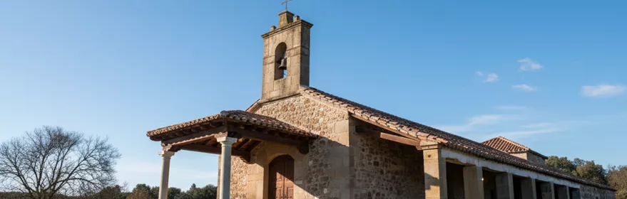 Una ermita rural de piedra con un largo soportal lateral sostenido por columnas, situada en un campo abierto bajo un cielo azul.
