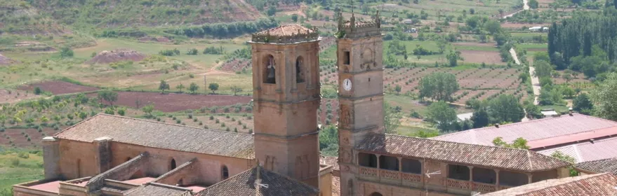 Vista aérea de un conjunto urbano con iglesia de piedra y tejados de teja rodeados de campos agrícolas.