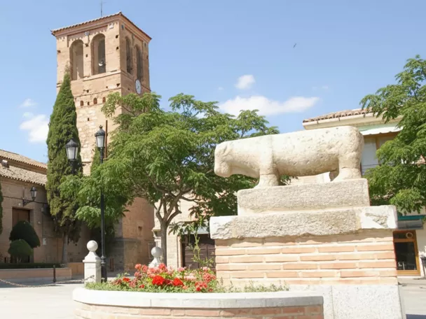 Fachada lateral de una iglesia de piedra y ladrillo con una torre campanario, árboles ornamentales y una escultura zoomorfa en primer plano.