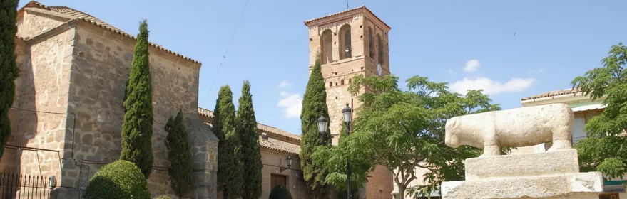 Fachada lateral de una iglesia de piedra y ladrillo con una torre campanario, árboles ornamentales y una escultura zoomorfa en primer plano.