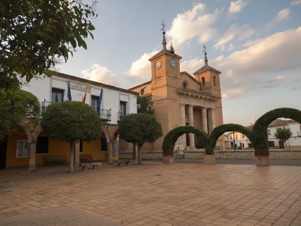 Una plaza de pueblo pavimentada con una iglesia de ladrillo al fondo, un edificio municipal con balcones a la izquierda y varios árboles de copa redonda y arcos vegetales podados.