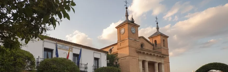 Una plaza de pueblo pavimentada con una iglesia de ladrillo al fondo, un edificio municipal con balcones a la izquierda y varios árboles de copa redonda y arcos vegetales podados.