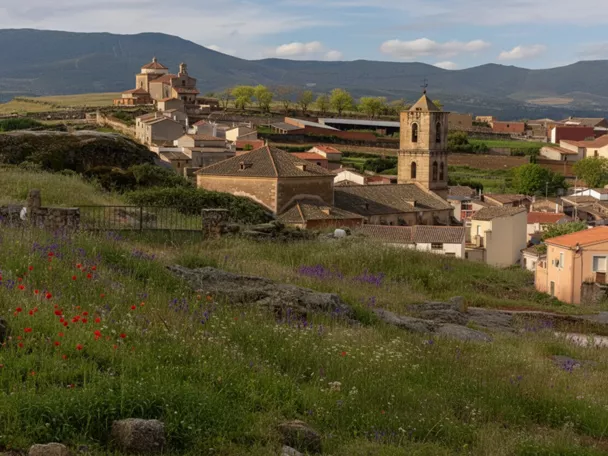 Paisaje rural con un pueblo de casas bajas y tejados de teja. En primer plano, un prado verde con flores silvestres rojas y moradas; al fondo, una iglesia con torre cuadrada y montañas bajo un cielo nublado.