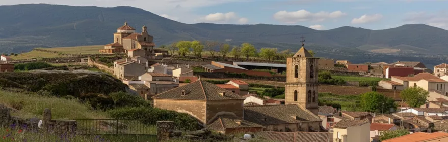 Paisaje rural con un pueblo de casas bajas y tejados de teja. En primer plano, un prado verde con flores silvestres rojas y moradas; al fondo, una iglesia con torre cuadrada y montañas bajo un cielo nublado.