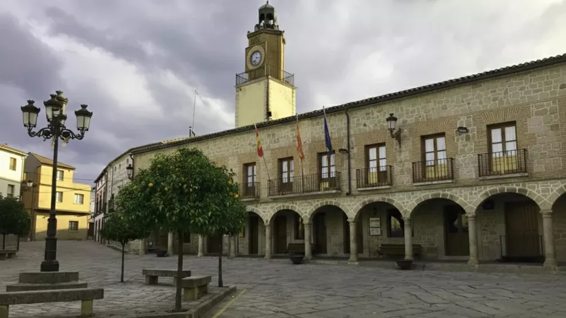Plaza mayor empedrada con un edificio consistorial de piedra que presenta una arquería en la planta baja y una torre de reloj amarilla.