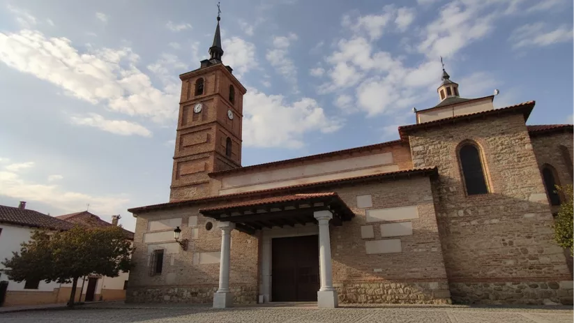 Fachada lateral de una iglesia de ladrillo y piedra con una torre campanario alta que tiene dos relojes y un chapitel oscuro.