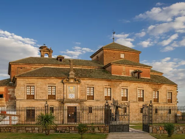 Un edificio histórico de piedra y ladrillo, con múltiples ventanas y un pequeño campanario, se encuentra detrás de una ornamentada valla de hierro forjado bajo un cielo nublado.