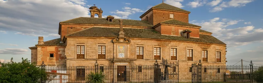 Un edificio histórico de piedra y ladrillo, con múltiples ventanas y un pequeño campanario, se encuentra detrás de una ornamentada valla de hierro forjado bajo un cielo nublado.