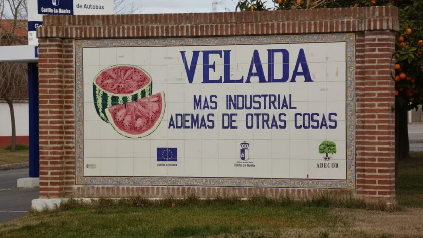 Un letrero de cerámica montado en una pared de ladrillo, con texto azul y una imagen de sandías, rodeado de árboles frutales y vegetación.