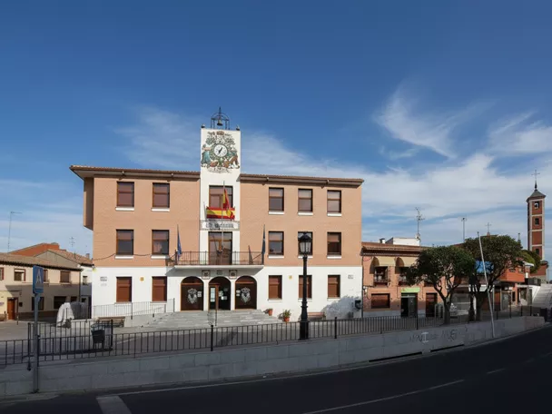 Vista amplia de una plaza de pueblo con un edificio administrativo principal en primer plano y una torre de iglesia visible a lo lejos.