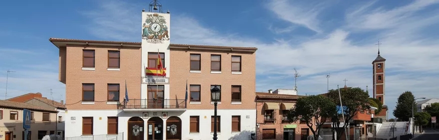Vista amplia de una plaza de pueblo con un edificio administrativo principal en primer plano y una torre de iglesia visible a lo lejos.