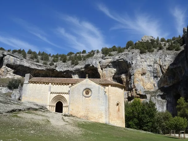 Ermita de piedra de estilo románico situada en la base de un imponente desfiladero rocoso, rodeada de vegetación y bajo un cielo azul despejado