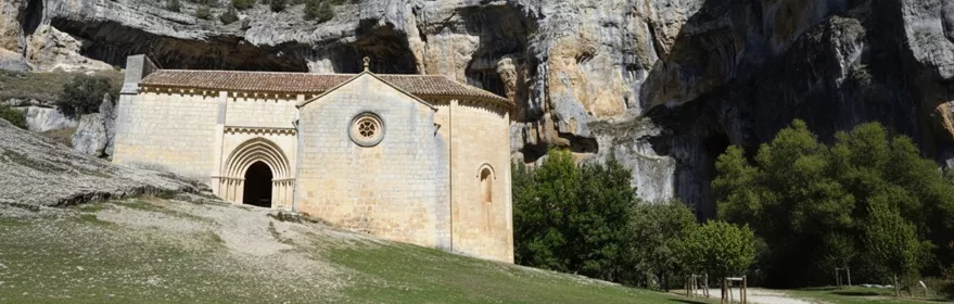 Ermita de piedra de estilo románico situada en la base de un imponente desfiladero rocoso, rodeada de vegetación y bajo un cielo azul despejado